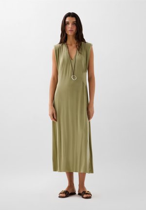 Abito lungo senza maniche di un verde oliva con scollatura a V profonda e arricciature laterali, abbinato a sandali marroni con un accento metallico decorativo.