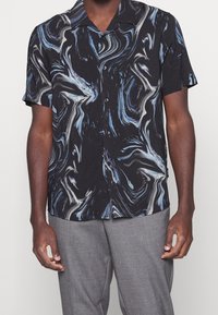 Uomo che indossa una camicia nera a maniche corte con abbottonatura e modelli astratti blu e bianchi, abbinata a pantaloni grigi.