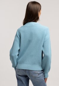 Jasnoniebieski sweter z dzianiny o teksturowym wzorze, z opadającymi ramionami i ściągaczami. Połączony z jasnoniebieskimi dżinsami.