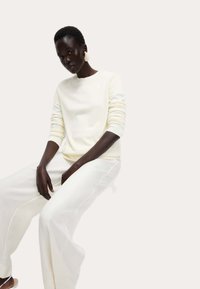 Jasna kremowa sweter z prążkowanymi mankietami i dołem, zestawiona z luźnymi białymi spodniami, o gładkiej, miękkiej teksturze. Minimalistyczny design.