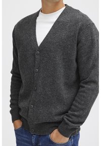 Cardigan de lana gris con cuello en V, seis botones, puños acanalados y un acabado ligeramente texturizado. Llevado sobre una camiseta blanca.