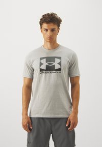 Siva pamučna majica s istaknutim crnim Under Armour logotipom u sredini. Kratki rukavi i klasičan dizajn s okruglim izrezom.