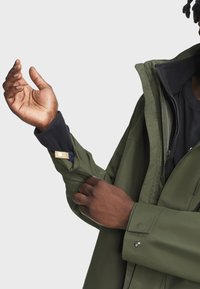 Zelená voděodolná bunda se zapínáním na zip, rukávy s fleecovou podšívkou, patentky a měkkou texturou, částečně zobrazená na zdvižené ruce.