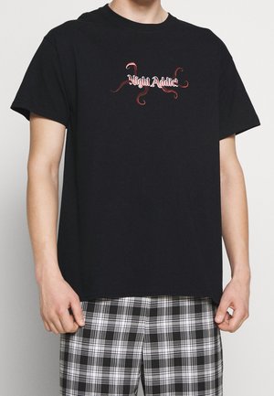 T-shirt in cotone nero con una grafica "Night Addict" rossa e bianca. Il modello lo indossa con pantaloni corti in plaid bianco e nero.