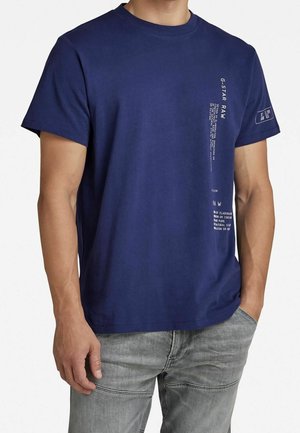 Mørkeblå bomuld T-shirt med rund halsudskæring, korte ærmer og trykte tekstdetaljer på fronten og ærmet. Bruges sammen med grå jeans.