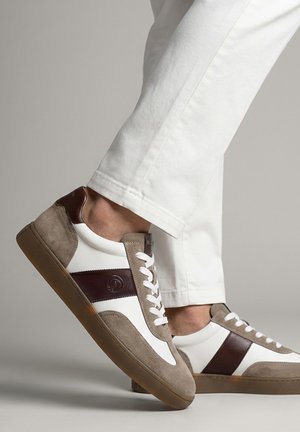 Person wearing white pants and white sneakers with brown suede and leather accents stepping forward indoors.