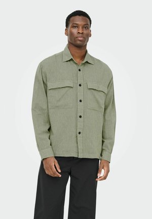 Un homme se tient debout, portant une chemise à boutons vert clair avec des poches sur la poitrine et un pantalon noir, devant un fond clair uni.