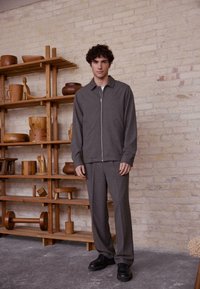 Grijze ritsjack en bijpassende broek van een zachte stof. De outfit heeft een kraag en een recht gesneden ontwerp, gecombineerd met zwarte schoenen.