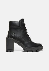 Bota de tobillo de cuero negro con un tacón grueso, que presenta un diseño de cordones, un cuello acolchado y una suela de goma tipo lug para mayor tracción. Se puede ver el grabado del logo.