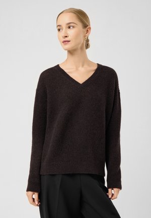 Woman with blonde hair in a bun wearing small hoop earrings, a dark brown V-neck sweater, and black pants, looking to her left.