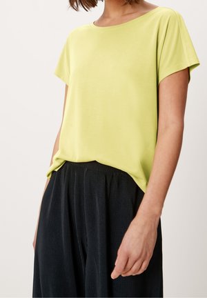 Frau trägt eine kurzärmelige, zitronengelbe Bluse, die in hochtaillierte, schwarze, plissierte, weit geschnittene Hosen gesteckt ist, vor einem schlichten Hintergrund.