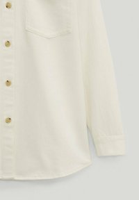 Chemise crème à manches longues avec boutons à l'avant, poche poitrine et poignets boutonnés sur un fond uni clair.