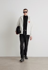 Felpa bianca con accenti rossi, dolcevita nera, jeans neri e stivali neri, accessoriata con occhiali da sole e una borsa nera.
