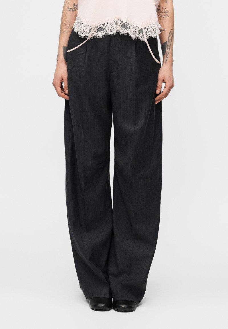 Άτομο που φοράει ψηλόμεσα, φαρδιά γκρι σκούρα παντελόνια με μαύρα παπούτσια και ένα ανοιχτό ροζ τοπ με δαντέλα, στέκεται μπροστά σε άσπρο φόντο.