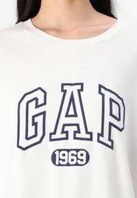 Balta kokvilnas T-kreklu ar spilgtu tumši zilu uzrakstu "GAP", zem kura mazā aplī ir uzraksts "1969". Ar apkakles dizainu.