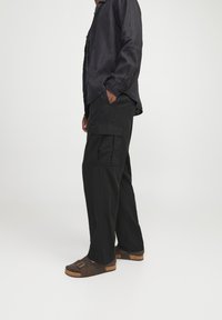 Pantalones cargo negros, holgados, con bolsillos laterales, combinados con una camisa de botones oscura y sandalias marrones con doble hebilla.