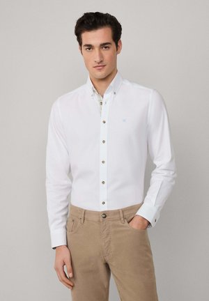 White button-up shirt with a collar, featuring brown buttons and a light blue logo. Worn with khaki pants. Plain texture, tailored fit.