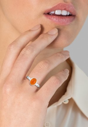 Sølvring med en oval orange sten, enkel ringskabelon. Hånd der blidt rører ansigtet, neutral baggrund, blød hudtekstur synlig.