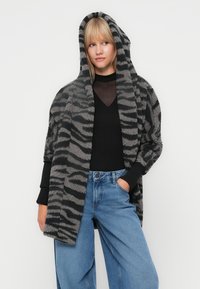 Veste à capuche surdimensionnée rayée gris et noir en tissu moelleux et texturé ; portée sur un haut noir et un jean large bleu clair.