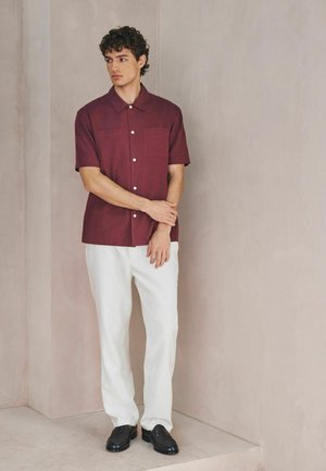 Cămașă bordo cu mâneci scurte, cu nasturi în față și două buzunare la piept, asortată cu pantaloni albi și mocasini negri, stând în fața unui perete neutru.