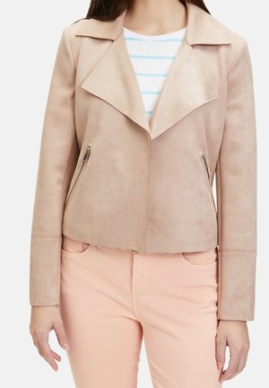 Femme portant une veste en daim beige clair avec de larges revers, des poches zippées, par-dessus une chemise blanche et bleue à rayures et un pantalon rose clair.