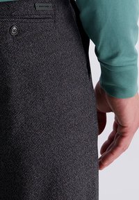 Gros plan d'une personne portant un pantalon texturé sombre avec une poche arrière à boutons et une chemise à manches longues turquoise, la main détendue sur le côté.