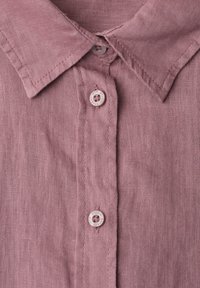 Gros plan du col d’une chemise en lin rose poudré avec trois boutons assortis et des coutures visibles le long des bords et de la patte de boutonnage.