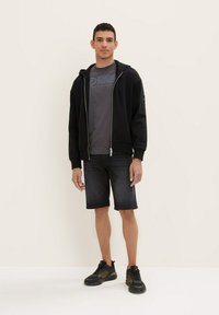 Haut à zip noir sur un T-shirt gris, associé à un short en denim foncé et des chaussures de sport noires. Design simple avec des détails fonctionnels.
