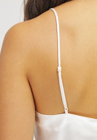 Camicetta in satin bianco con spalline sottili e chiusura sul retro. Il tessuto ha una texture liscia con un leggero luccichio.