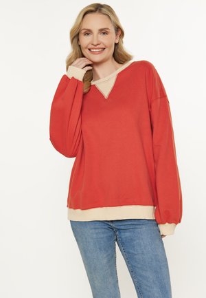 Rød sweatshirt med V-hals og beige manchetter, med lange ærmer og en afslappet pasform, parret med blå jeans.