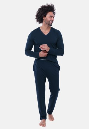 Man with curly hair wearing navy blue long-sleeve top and pants, standing barefoot, smiling, and looking to the side against a plain background.