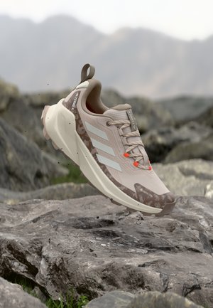 Beiger Trailrunning-Schuh mit strukturiertem Material, drei weißen Streifen, orangefarbenen Akzenten und robustem Sohlen-Design; verfügt über eine verstärkte Ferse und ein Gore-Tex-Logo.
