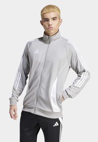 Grå atletisk jakke med lynlås, hvide detaljer og tredobbelt stribe langs ærmerne. Lavet af glat, let vævning.