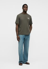 Άνδρας που στέκεται, φοράει κοντομάνικο T-shirt σε ελαιόχρωμο πράσινο με λευκό κείμενο, ανοιχτόχρωμα τζιν παντελόνια φαρδιάς γραμμής και καφέ σανδάλια ανοιχτά στη μύτη, σε λευκό φόντο.