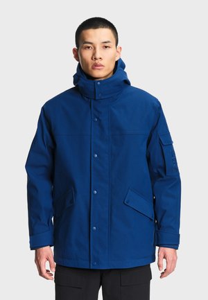 Homme portant une veste imperméable à capuche bleue avec boutons à l'avant et poches à rabat, debout devant un fond uni.