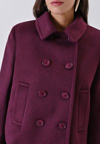Cappotto doppiopetto viola realizzato in tessuto strutturato, con grandi bottoni, ampio colletto e tasche laterali. Progettato per una vestibilità aderente.