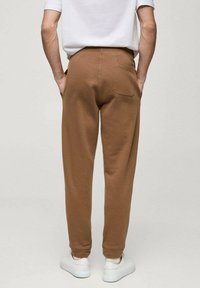 Pantalonii de trening maro au o croială lejeră, talie elastică, un singur buzunar la spate și sunt asortați cu adidași albi.