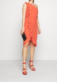 Koralowa sukienka na jedno ramię z falbaną, o długości do kolan i taliowana w pasie. Dobierz do niej czerwone sandały na wysokim obcasie z paskami oraz czarną kopertówkę.