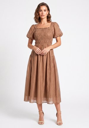 Donna che indossa un vestito midi marrone con maniche a sbuffo, corpetto arricciato e tacchi beige con cinturino alla caviglia, in piedi davanti a uno sfondo bianco semplice.