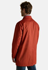 Raudonas, vandeniui atsparus paltas su paprastu dizainu, turintis tiesų kirpimą, užsegamus rankovės galus ir apykaklę. Galinis vaizdas rodo minimalų detalizavimą.