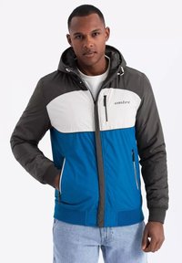 Giacca imbottita con corpo blu, parte superiore anteriore bianca e maniche grigie. Presenta un cappuccio, tasche con zip e polsini e fondo a costine.