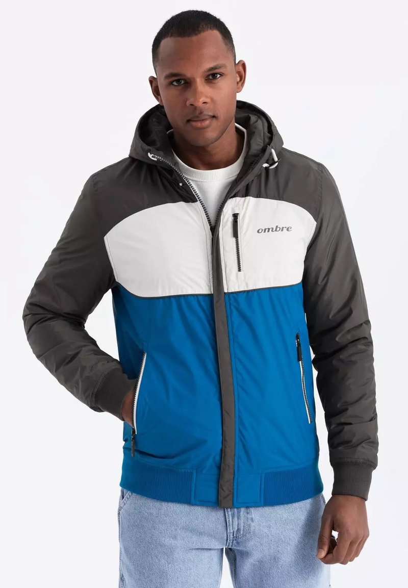 Giacca imbottita con corpo blu, parte superiore anteriore bianca e maniche grigie. Presenta un cappuccio, tasche con zip e polsini e fondo a costine.