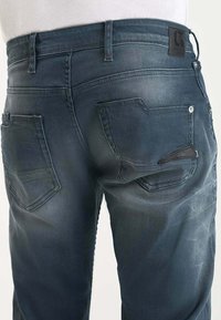 Vaqueros de denim azul oscuro con detalles desvanecidos, vista trasera mostrando dos bolsillos traseros, pasadores de cinturón y una etiqueta de cuero de la marca en la cinturilla.