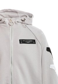 Sudadera con capucha gris claro con cremallera, cuello alto, cordones, parche rectangular negro en el pecho y parches de alas en blanco y negro en la manga.