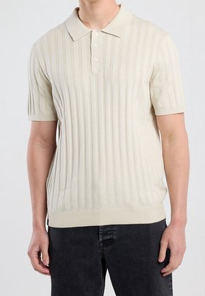 Mężczyzna w kremowej, prążkowanej koszulce polo z krótkim rękawem w połączeniu z czarnymi dżinsami, stojący na jednolitym tle.