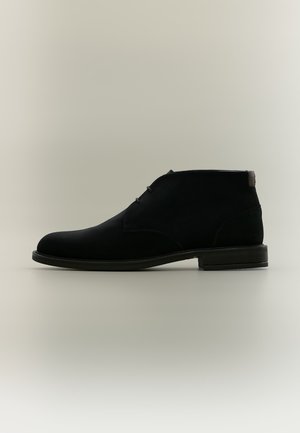 Bottine en daim noire avec des lacets minimalistes, bout arrondi et talon bas empilé sur un fond uni clair.