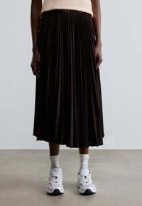 Jupe midi plissée noire avec une ceinture élastique arborant un détail de marque. Associée à des baskets blanches et des chaussettes montant jusqu'à la cheville.