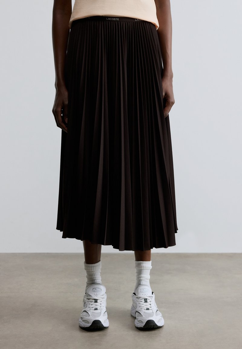 Jupe midi plissée noire avec une ceinture élastique arborant un détail de marque. Associée à des baskets blanches et des chaussettes montant jusqu'à la cheville.