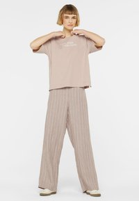 Locker sitzendes, blassrosa T-Shirt mit dem Text "LOVE OVER EVERYTHING", kombiniert mit beigen, gestreiften, weit geschnittenen Hosen und hellen Schuhen.