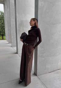 Vrouw in een bruine coltrui en wijde gestreepte broek leunt tegen een betonnen pilaar en houdt een zwarte clutch vast in een moderne buitenarchitectuur omgeving.